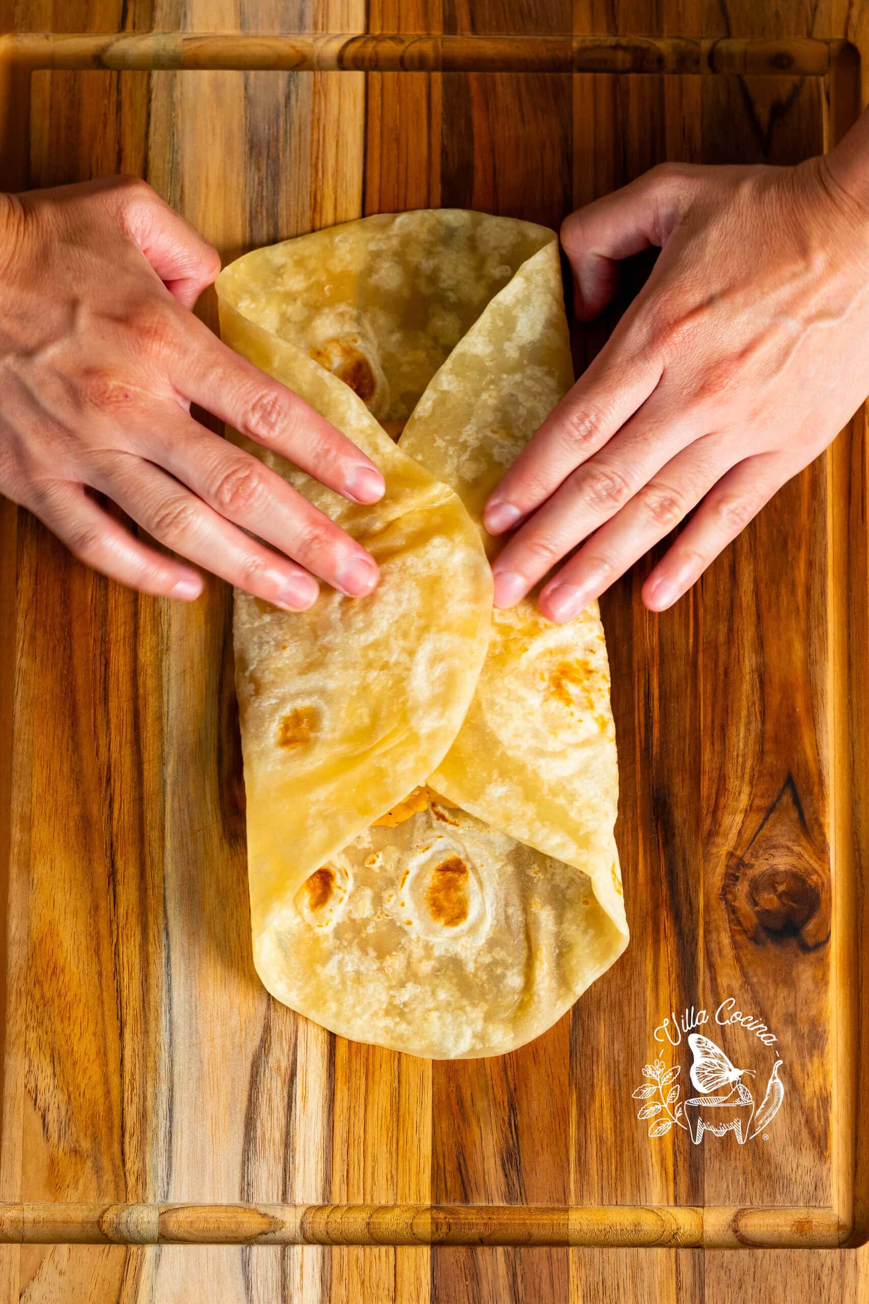 Hands folding a tortilla to make a bean and cheese burrito on a wooden surface.