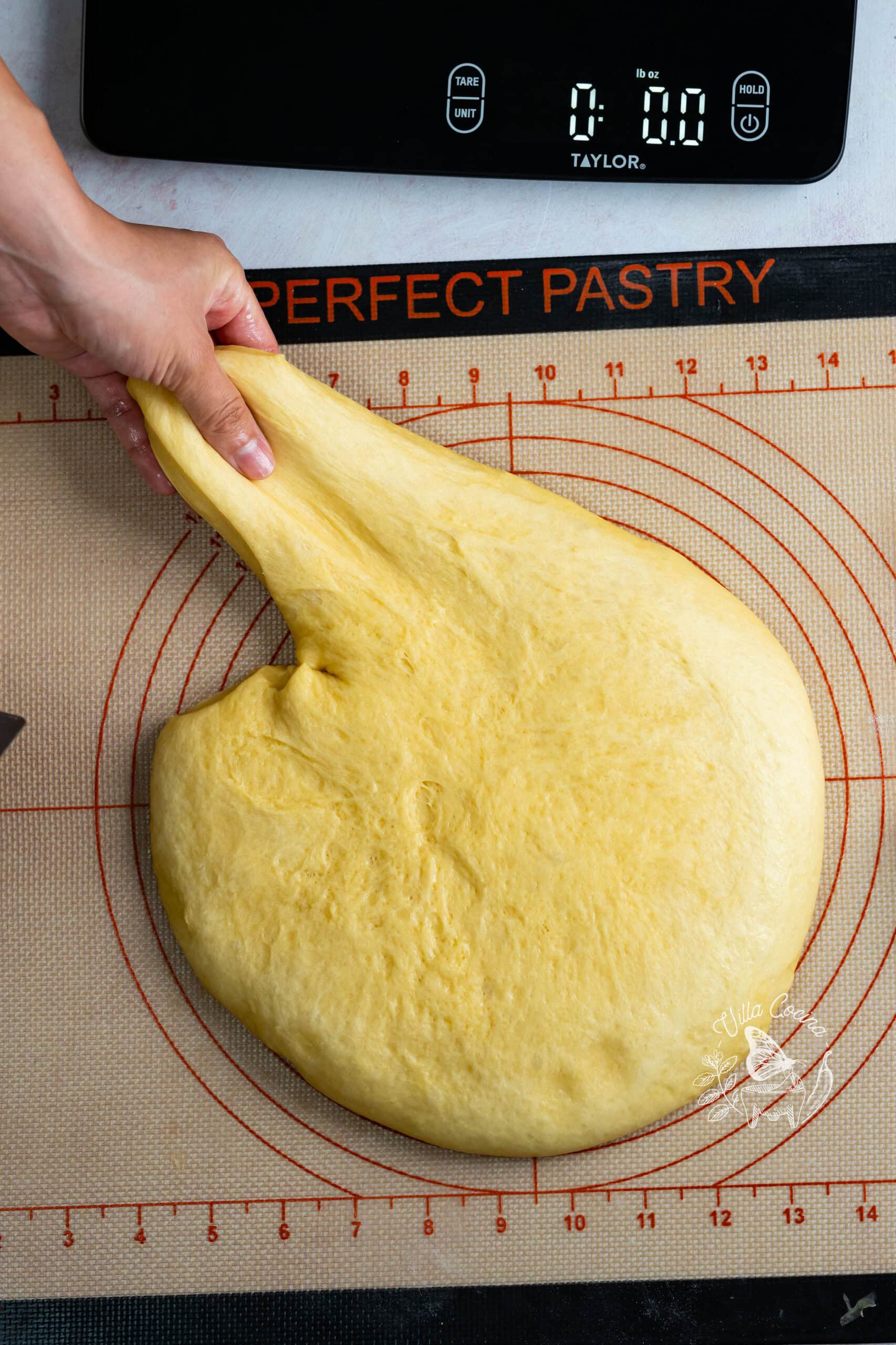Hand stretching risen concha dough on a silicone mat beside a kitchen scale.
