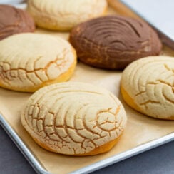 Close view of homemade Mexican conchas with vanilla and chocolate sugar toppings, showing seashell patterns and golden bread on a baking sheet.